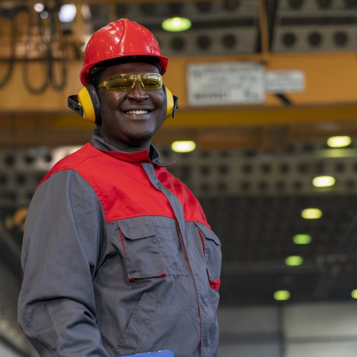 Waist Up Portrait Of Black Industrial Worker In Red Helmet, Yellow Safety Goggles, Noise Reduction Earmuffs And Work Uniform In A Factory.
