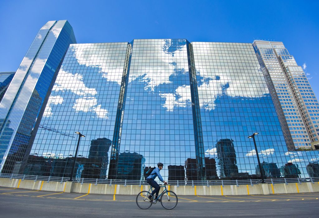 A male bicycle commuter rides his bike through a parking lot on his way to work in a big city. He rides a single-speed or fixed-gear bicycle with just a front brake. He wears a cycling helmet and carries a bicycle messenger-style backpack. He rides past the office buildings that reflect the clouds in the sky.