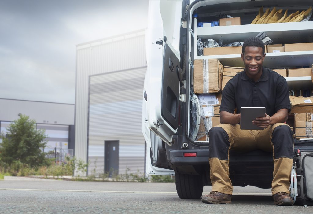 A service engineer sat at the back of his van using a digital tablet outside factory buildings