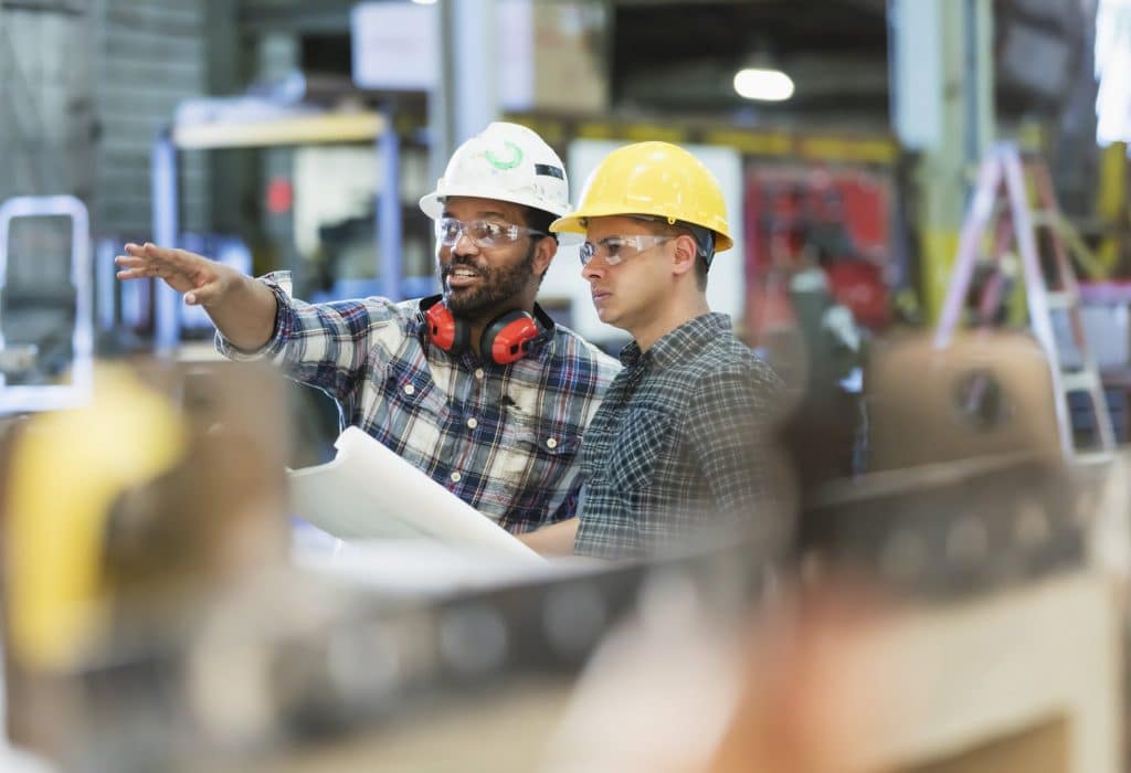 Two multi-ethnic workers in their 30s talking in a metal fabrication plant wearing hardhats and protective eyewear. The man pointing is African-American and his coworker is Hispanic.